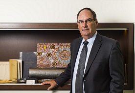 Deputy Electoral Commissioner David Gully is a white male with glasses, in his mid to late fifties, and is wearing a black suit and tie and a white shirt. He is standing beside a cabinet housing an aboriginal dot painting and other historical electoral items.